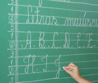 Professora escrevendo letras maiúsculas no quadro verde, com as pautas numeradas 1, 2 e 3 desenhadas a giz